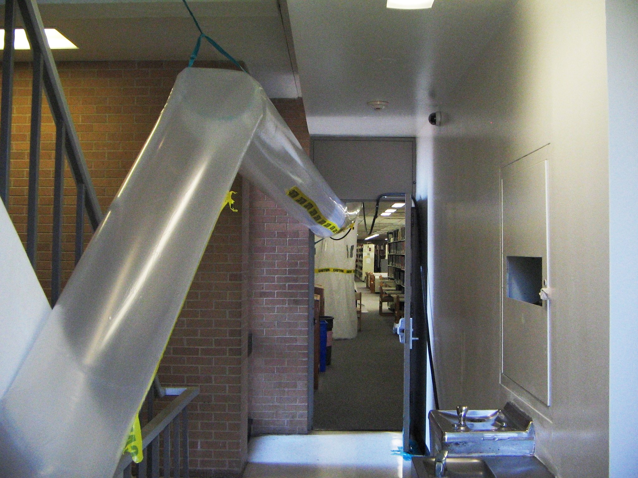 A large temporary clear plastic air duct runs through an interior hallway beside caution tape and an enclosed work area.